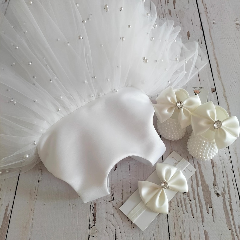 May include: White tulle baby dress with pearl embellishments, a matching headband with two satin bows, and a pair of white satin baby shoes with pearl embellishments and satin bows.