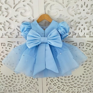 May include: A pale blue baby dress featuring a large bow and short puff sleeves. The dress has a tulle skirt decorated with small, sparkling embellishments. It hangs on a wooden hanger against a white, ornate background.