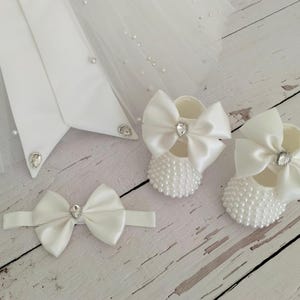 May include: White satin baby shoes with pearl embellishments and a large white satin bow. The shoes have a rhinestone in the center of the bow.