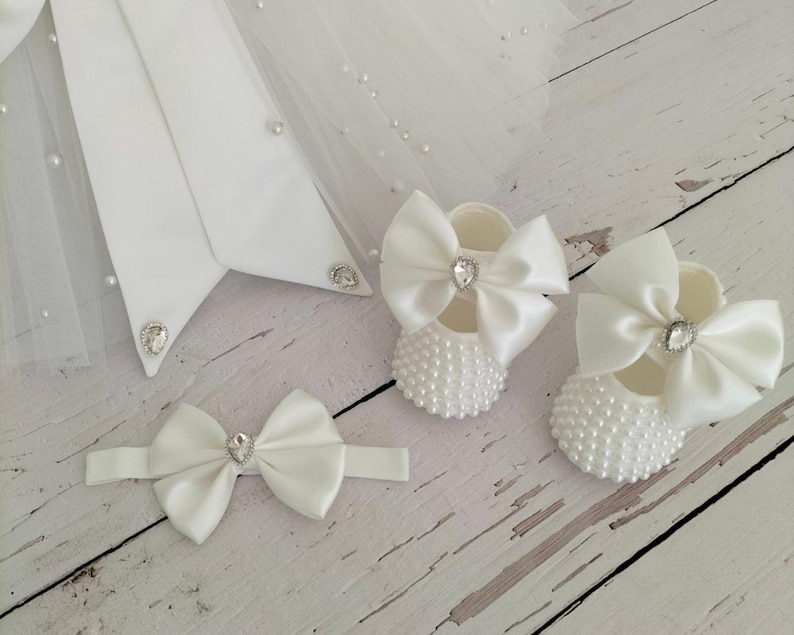 May include: White satin baby shoes with pearl embellishments and a large white satin bow. The shoes have a rhinestone in the center of the bow.