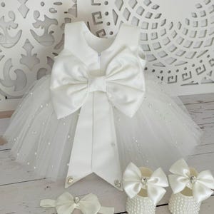 May include: White tulle baby dress with a large satin bow and pearl embellishments. The dress comes with a matching headband and shoes.