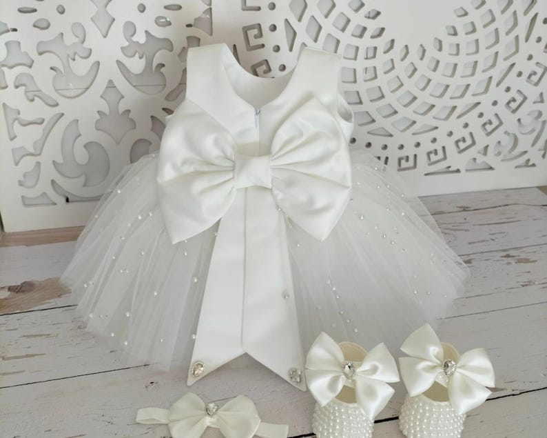 May include: White tulle baby dress with a large satin bow and pearl embellishments. The dress comes with a matching headband and shoes.