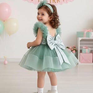 May include: A little girl in a mint green tulle dress with a large bow on the back. The dress has ruffled sleeves and a matching headband. Balloons in pastel colors and wrapped gifts are in the background.