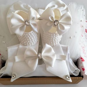 May include: White baby shoes with pearl embellishments and satin bows, a white headband with a satin bow, and a white hat with a satin bow. The items are arranged in a gift box with a white and pink patterned paper.