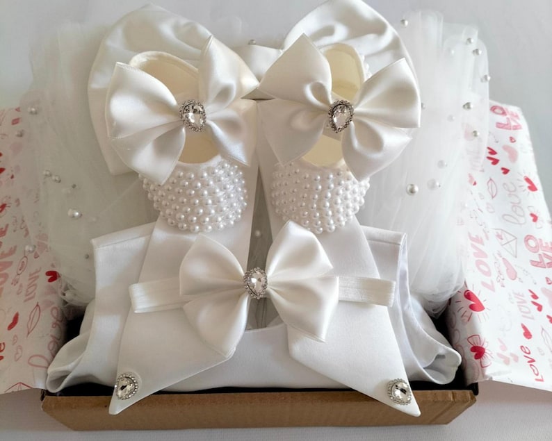 May include: White baby shoes with pearl embellishments and satin bows, a white headband with a satin bow, and a white hat with a satin bow. The items are arranged in a gift box with a white and pink patterned paper.