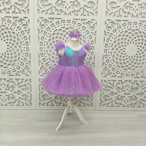 May include: A lavender tutu dress with a sequined bodice and tulle skirt, adorned with small white pearls. The dress features matching lavender tulle sleeves and a headband with a bow. The dress is displayed on a white mannequin.