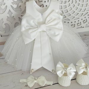 May include: White tulle baby dress with a large satin bow on the back. The dress is decorated with small white pearls. The set includes a matching headband and a pair of white shoes with pearl embellishments.