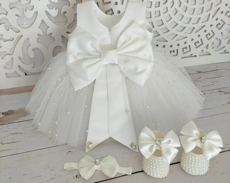 May include: White tulle baby dress with a large satin bow on the back. The dress is decorated with small white pearls. The set includes a matching headband and a pair of white shoes with pearl embellishments.