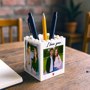 May include: A white block-style pen holder with a photo of a couple, holding pens and a pencil. The words "I love you" are written on the side. The pen holder is on a wooden table next to an open notebook.