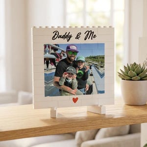 Peut inclure: Cadre photo blanc avec le texte "Daddy & Me" au-dessus d'une photo d'un homme et d'un enfant. Le cadre a un cœur rouge en bas et est exposé sur une étagère en bois avec d'autres objets décoratifs.