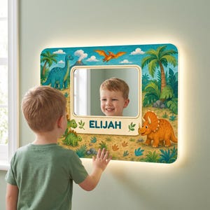 May include: A rectangular wall mirror featuring a colorful dinosaur-themed border with illustrations of a blue long-necked dinosaur, an orange triceratops, and a flying pterodactyl. The name ELIJAH is printed in blue text below the mirror.