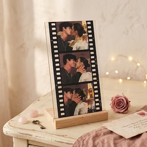 May include: A clear acrylic photo display in the style of a film strip, showing three images of a couple kissing. It is mounted on a natural wood base. A vintage key and a rose are on the table.