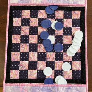 Può includere: Una scacchiera trapuntata con bordo nero e accenti rosa e viola. Le caselle della scacchiera si alternano tra un motivo a pois blu scuro e un disegno a piume rosa e gialle. Le pedine blu e bianche sono sparse sulla scacchiera.