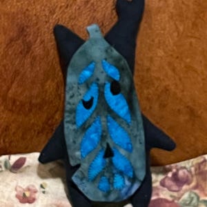 May include: A handmade plush doll shaped like a creature with a leaf-like face. The doll is mainly navy blue with a teal and blue leaf design on the front. The leaf has blue leaf-shaped accents and black eyes.