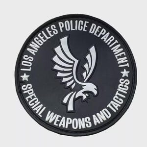 May include: A circular black patch with white text reading "LOS ANGELES POLICE DEPARTMENT SPECIAL WEAPONS AND TACTICS". The patch features a white eagle emblem in the centre.