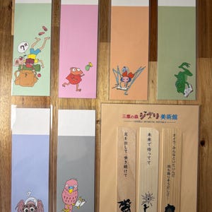 Puede incluir: Un conjunto de marcapáginas coloridos con ilustraciones de dibujos animados. Los marcapáginas tienen una sección superior blanca y una sección inferior en varios colores, incluyendo verde, rosa y azul. Algunos marcapáginas tienen texto japonés.