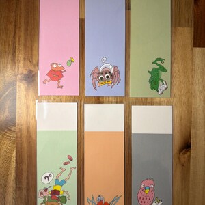May include: A collection of six illustrated bookmarks. Each bookmark has a different pastel background and features a cartoon character, such as a frog or spider. The top section of each bookmark is white.