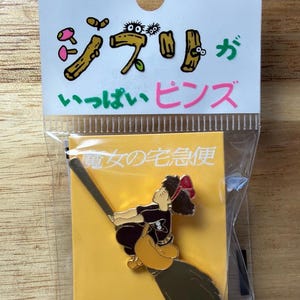 May include: A pin featuring a character from the animated film "Kiki's Delivery Service" riding a broom. The pin is packaged with a header card that has Japanese text and cartoon illustrations. The pin is in a clear plastic bag.