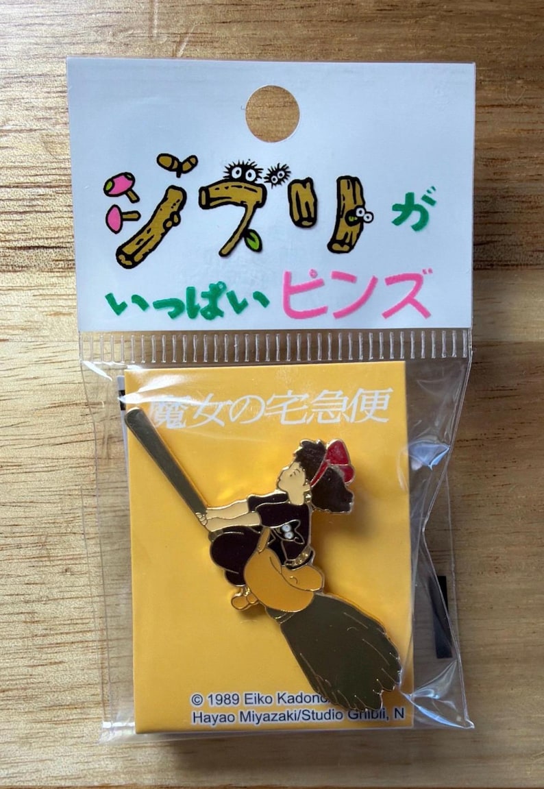 May include: A pin featuring a character from the animated film "Kiki's Delivery Service" riding a broom. The pin is packaged with a header card that has Japanese text and cartoon illustrations. The pin is in a clear plastic bag.