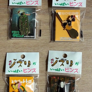 May include: Four Studio Ghibli pins in clear plastic packaging. Each pin features a different character or scene from popular animated films. The packaging includes Japanese text and the Studio Ghibli logo.