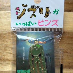 May include: A pin featuring a green robot from the movie "Castle in the Sky" with the Japanese text "Ghibli Pins" in pink and green. The pin is on a card with the Studio Ghibli logo.