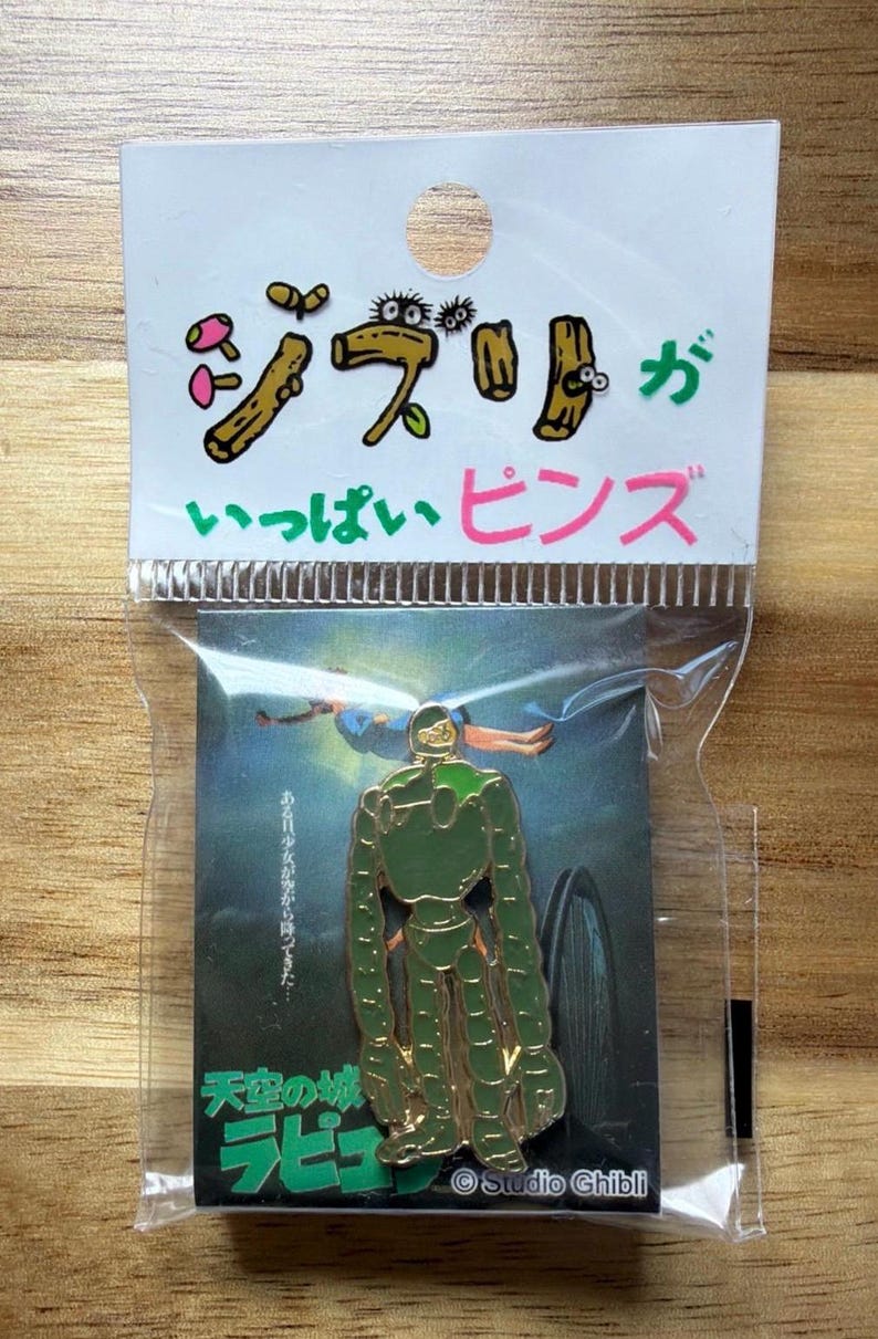May include: A pin featuring a green robot from the movie "Castle in the Sky" with the Japanese text "Ghibli Pins" in pink and green. The pin is on a card with the Studio Ghibli logo.
