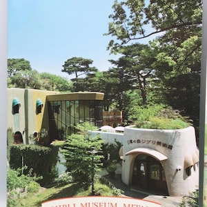 May include: A photograph of the Ghibli Museum in Mitaka, Japan, showcasing its unique architecture. The building features a whimsical design with rounded shapes, lush greenery, and the museum's name in a banner.