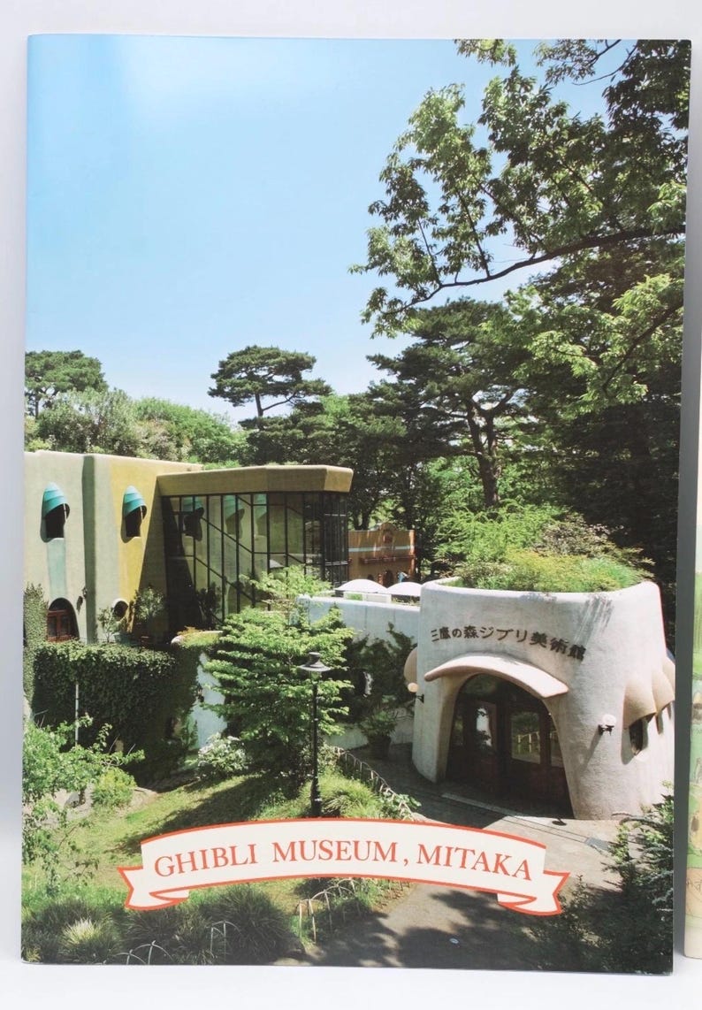 May include: A photograph of the Ghibli Museum in Mitaka, Japan, showcasing its unique architecture. The building features a whimsical design with rounded shapes, lush greenery, and the museum's name in a banner.