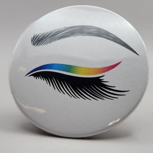 May include: A white button with a black and rainbow eye lash design. The eye lash is outlined in blue and has a rainbow colored stripe. The eyebrow is gray.