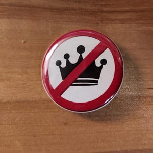 May include: A round button with a white background, a black crown, and a red slash through it. The button has a red border and is set against a wooden surface.