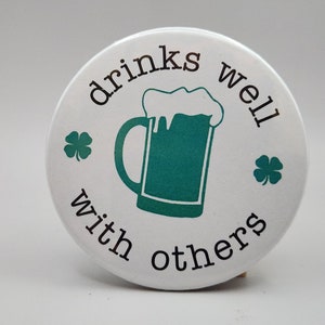 May include: White round button with the text "drinks well with others" in black. A green beer mug with a white foam top is in the center. There are two green shamrocks on either side of the text.