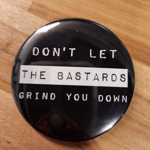 May include: A circular black pin-back button featuring white text that reads DON'T LET THE BASTARDS GRIND YOU DOWN. The text is arranged with the words DON'T LET on the top, THE BASTARDS on a white horizontal stripe, and GRIND YOU DOWN on the bottom.