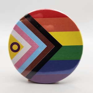 May include: A round button with a rainbow flag design. The flag includes the colors red, orange, yellow, green, blue, purple, brown, black, white, and pink. The flag also includes a yellow circle with a purple ring around it.