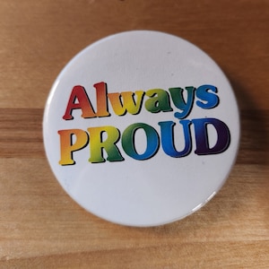 May include: A white button with the text "Always Proud" in rainbow colors.