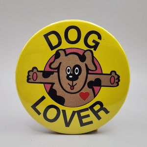 May include: Yellow button with the text "DOG LOVER" in black letters. A cartoon dog with brown fur, black spots, and a red heart on its chest is in the center of the button.