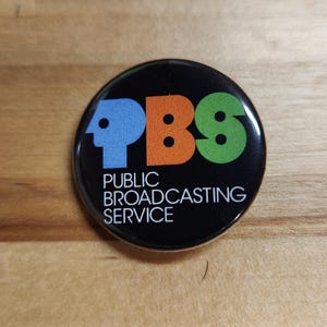 May include: A black round button with the PBS logo in blue, orange, and green. The text "PUBLIC BROADCASTING SERVICE" is printed in white below the logo. The button is on a wooden surface.
