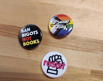 May include: Three round button pins with different messages and designs. The first pin is black with white text that says "Ban Bigots Not Books". The second pin is white with a pink fist and the word "resist" in pink. The third pin is black with a rainbow flag and the words "Proud Ally" in a rainbow font.