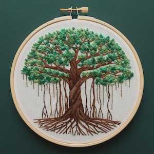 May include: An embroidered tree design in a wooden hoop. The tree features a brown trunk and roots, with green leaves and hanging roots. The embroidery is on a white fabric background.