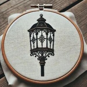 May include: An embroidered image of a vintage street lamp in black and white, displayed in a wooden embroidery hoop. The lamp has intricate detailing and is stitched on cream-colored fabric. The hoop rests on a white cloth.