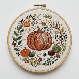 May include: A circular wooden embroidery hoop containing a fabric panel with a hand-stitched autumn scene. The design features a large central orange pumpkin with a white spiderweb, surrounded by smaller pumpkins, an owl, berries, and leaves.