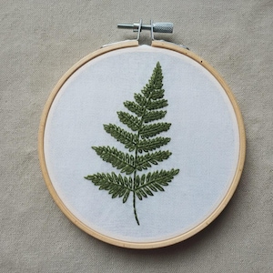 May include: A green fern leaf embroidered on white fabric, stretched within a wooden embroidery hoop. The fern's detailed stitching creates a textured appearance. The hoop is secured with a metal screw and sits on a neutral-toned background.