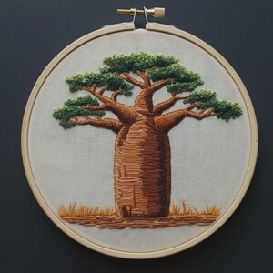 May include: A circular wooden embroidery hoop containing a hand-stitched baobab tree on off-white fabric. The tree features a thick, textured brown trunk with detailed branch structures and dense, layered green foliage, set against a patch of golden grass.