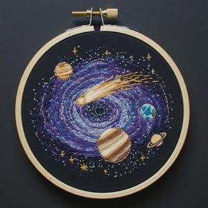 May include: A circular wooden embroidery hoop containing a hand-stitched space scene on black fabric. The design features a swirling purple and blue galaxy, a bright yellow comet, and several planets in shades of brown, blue, and tan with small stars.