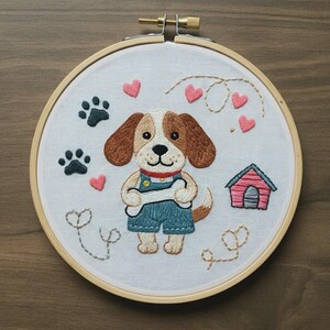 Puede incluir: Un bastidor de bordado circular de madera con tela blanca que presenta un diseño bordado a mano de un cachorro marrón y blanco con peto azul que sostiene un hueso blanco. El diseño incluye huellas de patas gris oscuro, corazones rosas y una caseta de perro rosa.