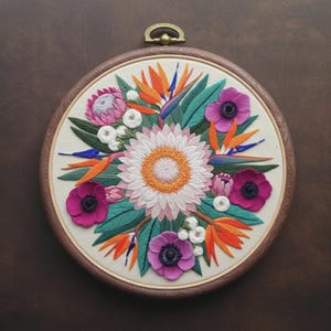 Può includere: Un telaio da ricamo circolare contenente un motivo floreale su tessuto bianco sporco. Il ricamo presenta un grande fiore centrale bianco con centro giallo, circondato da fiori uccello del paradiso arancioni, protee rosa e anemoni viola.