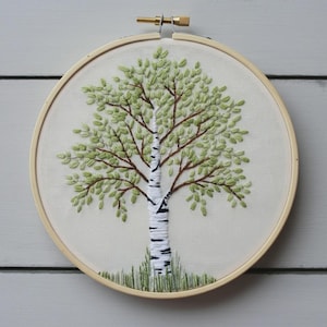 May include: Hand-stitched embroidery art of a birch tree in a wooden hoop. The tree features a white trunk with black markings, brown branches, and green leaves. Green grass is stitched at the base. The hoop is approximately 8 inches in diameter.
