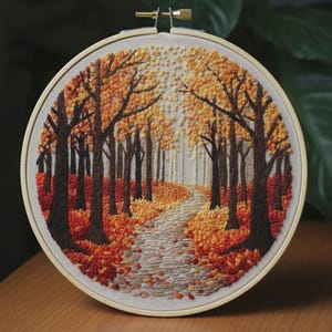 Puede incluir: Un bastidor de bordado circular que contiene un panel de tela con una escena forestal cosida a mano. El diseño presenta troncos de árboles de color marrón oscuro y un sendero sinuoso rodeado de follaje denso en tonos rojos, naranjas y amarillos.