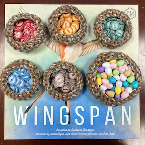 Könnte beinhalten: Das Brettspiel "Wingspan" mit sechs gehäkelten Nestern, die mit Spielsteinen gefüllt sind. Die Nester sind braun und beige, und die Spielsteine sind in verschiedenen Farben gehalten. Das Spielbrett ist mit dem Spieletitel und dem Namen des Designers sichtbar.