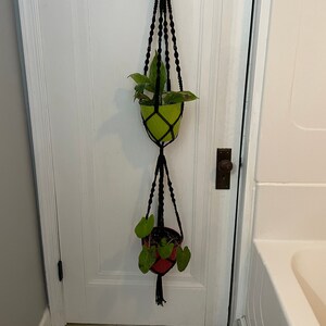 May include: A black macrame plant hanger with two colorful pots. The top pot is lime green, and the bottom pot is red. Both pots contain green plants. The hanger is suspended from a white door.