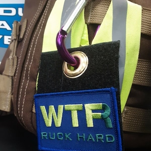 Op de afbeelding: Een zwarte patch met een blauwe rechthoek waarop in groen en geel "WTR RUCK HARD" staat. De patch is bevestigd aan een paarse karabijnhaak.
